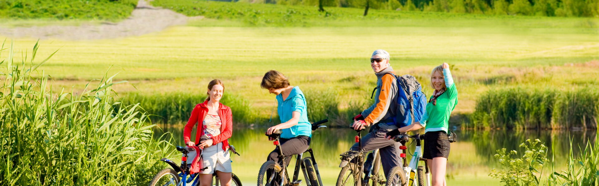 Aktiver Familienurlaub: Radtouren in der malerischen Landschaft um Crostau. Eine vierköpfige Familie steht mit Fahrrädern an einem See, umgeben von grünen Wiesen und bewaldeten Bergen unter blauem Himmel. Sie machen eine Pause während ihrer Fahrradtour.