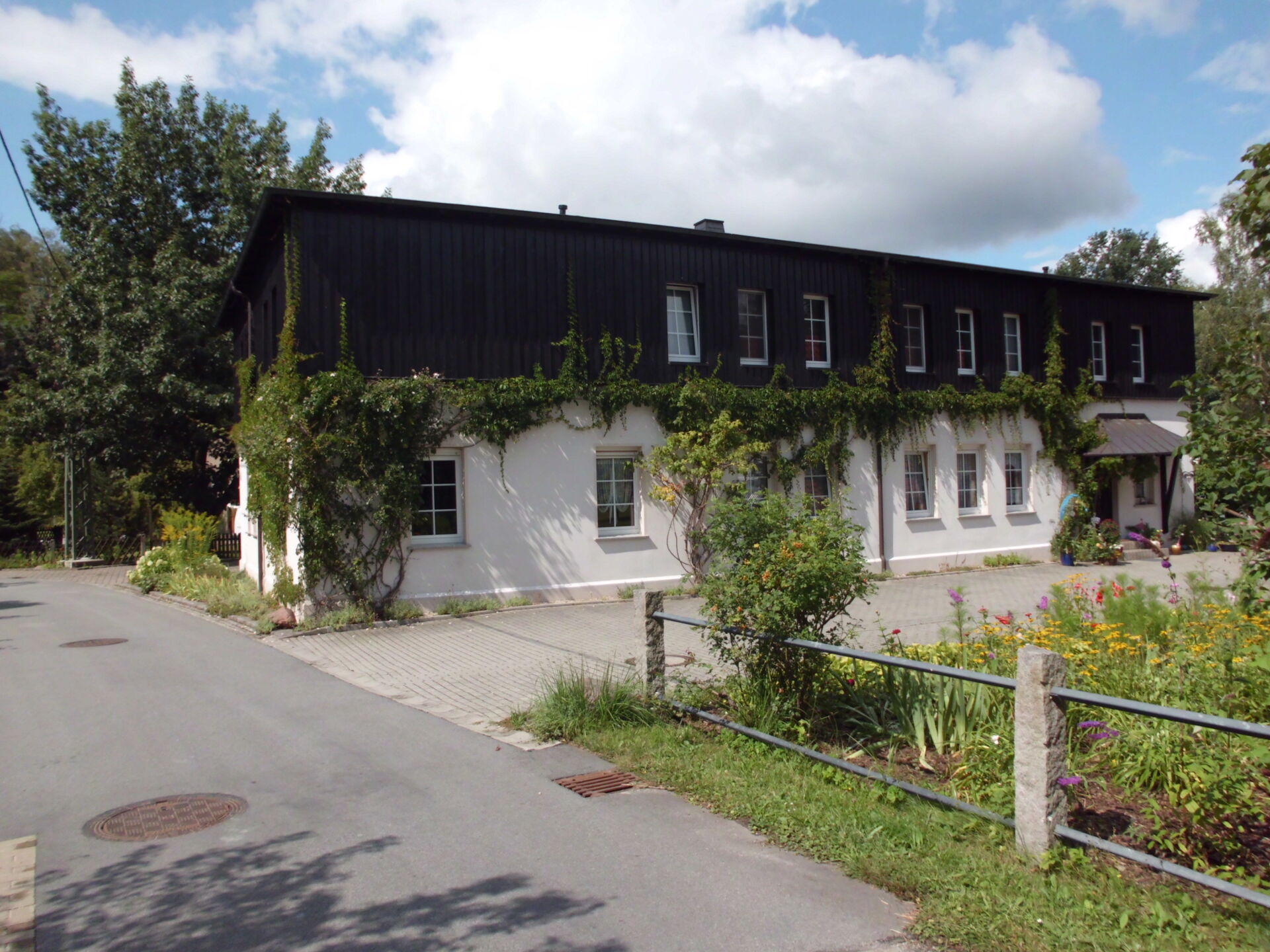 Ihre Unterkunft in Crostau: Herzlich willkommen in der Pension Adriaurlaub. Die Pension Adriaurlaub in Crostau: Ein charmantes, zweistöckiges Gebäude mit einer weiß verputzten unteren Etage und einer dunklen Holzfassade im oberen Bereich, teilweise bewachsen mit Rankpflanzen. Davor befindet sich ein kleiner Vorgarten mit Blumen u